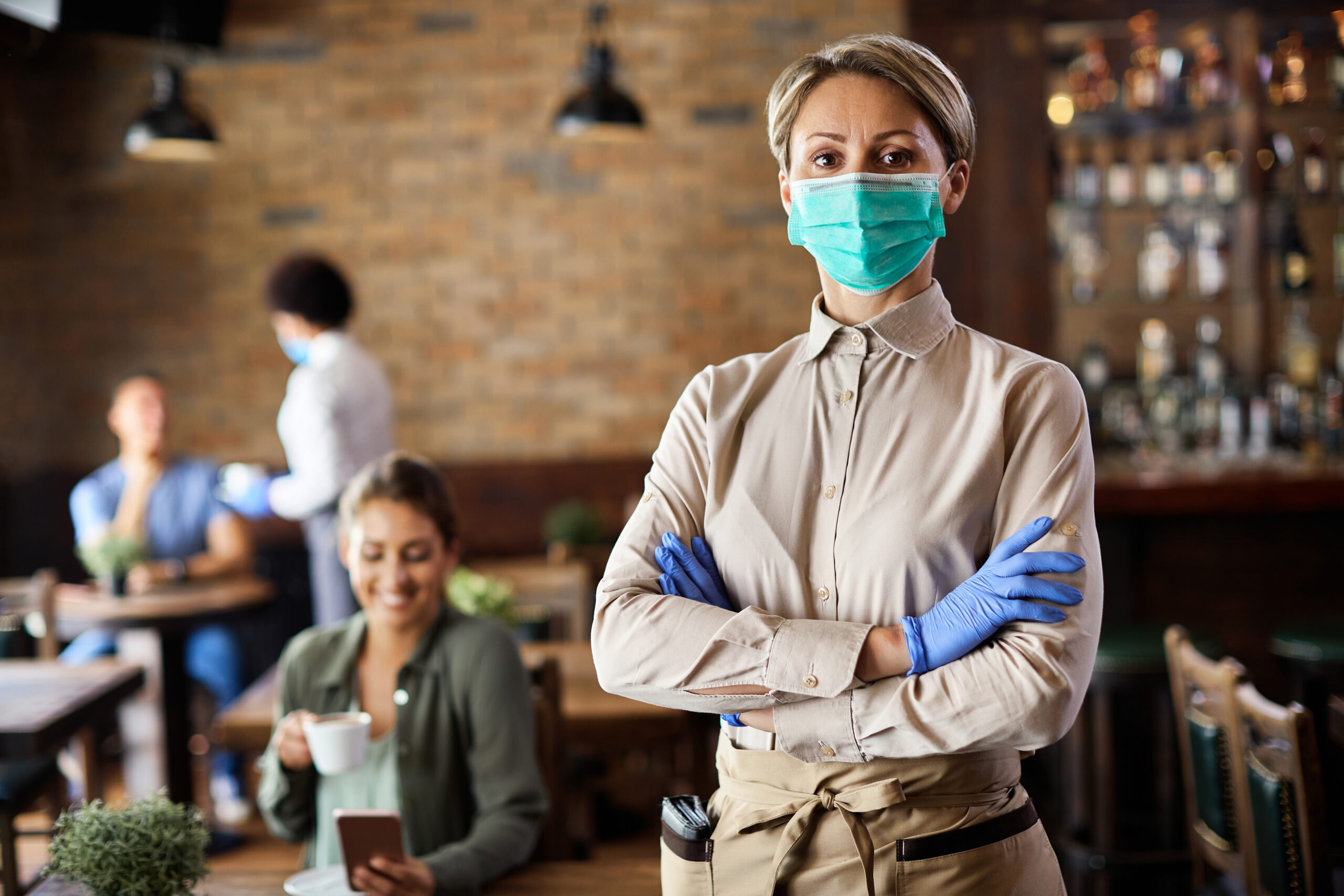Waitress standing with arms crossed while wearing protective face mask and working in a cafe during coronavirus epidemic.